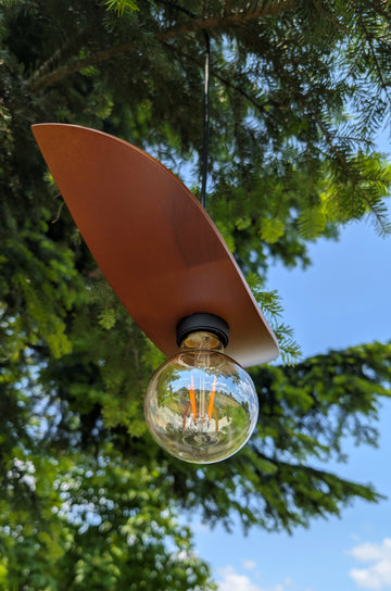 Surfboard-shaped lamp with a light bulb hanging outdoors against a tree and blue sky.