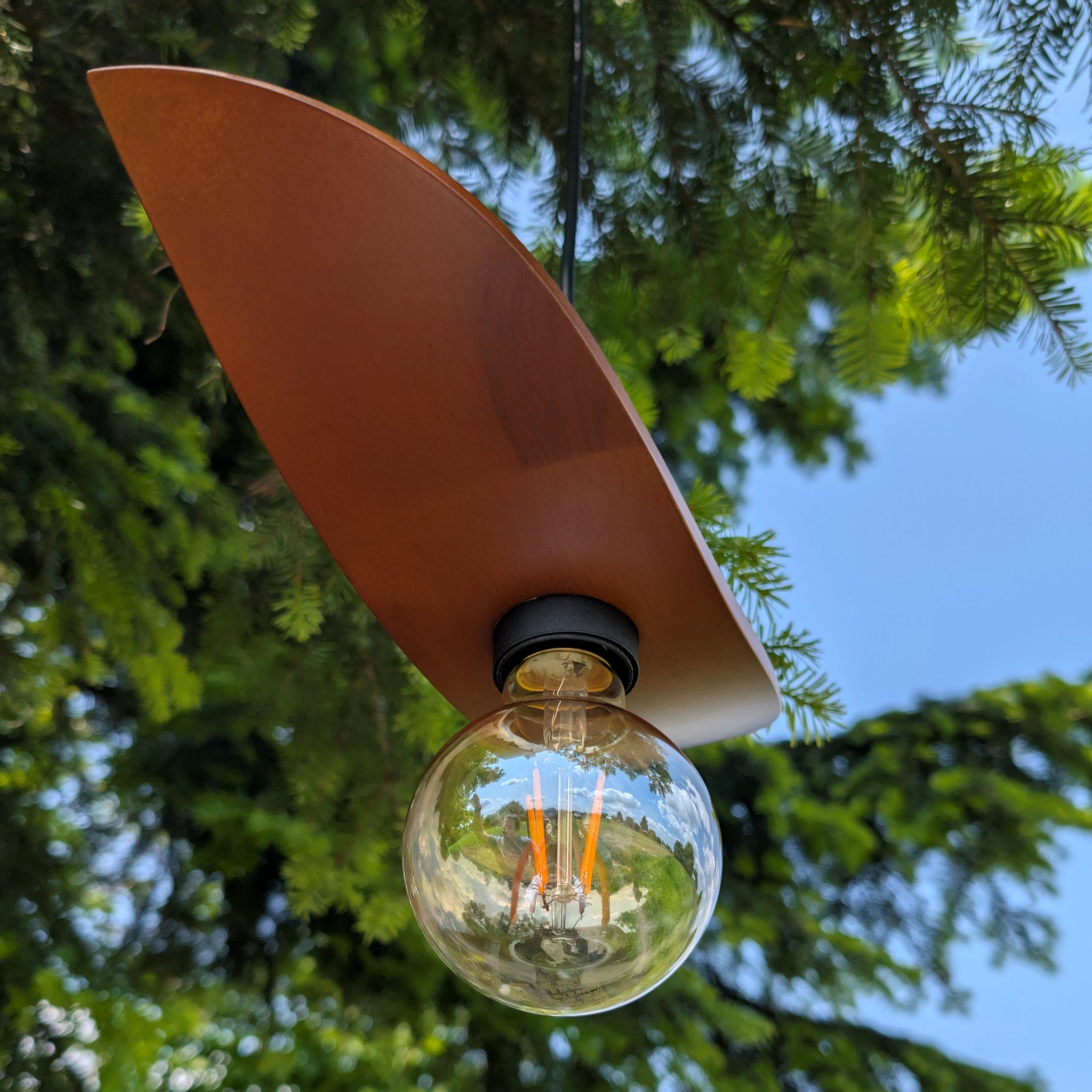 Surfboard-shaped lamp with a light bulb hanging outdoors against a tree and blue sky.